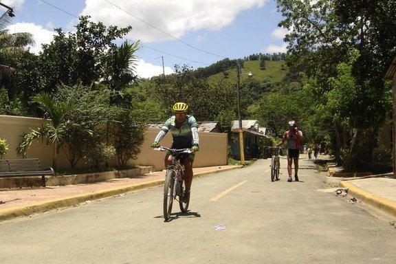 A scenic street with a cyclist wearing a yellow helmet riding a mountain bike, while another cyclist walks alongside their bike. Lush greenery lines the road, and hills are visible in the background, under a partly cloudy sky. Cordillera Central mountain bike trail.