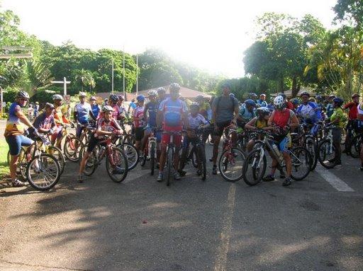A large group of cyclists, wearing colorful biking attire and helmets, is gathered at the starting point of a race or event. They are all on mountain bikes, standing on a paved road with greenery in the background. The scene is filled with anticipation and excitement as they prepare to begin their ride. Cordillera Central mountain bike trail.