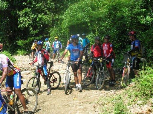 A group of mountain bikers gathered on a dirt path surrounded by greenery, some sitting on their bikes and others standing. They are wearing colorful cycling attire and helmets, engaged in conversation amidst a natural setting. Cordillera Central mountain bike trail.