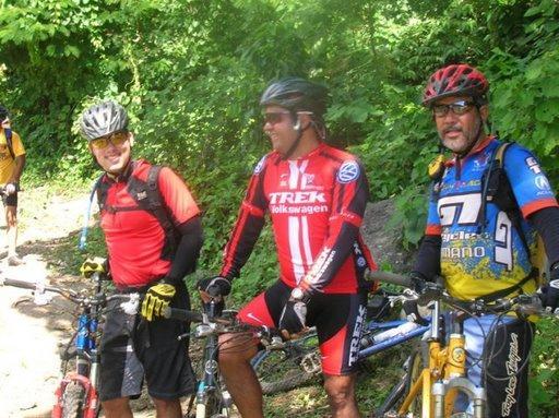 Three cyclists pause on a trail surrounded by greenery. They are wearing helmets and colorful cycling jerseys, each with unique designs. Two of the cyclists are standing next to their mountain bikes while the third leans slightly forward, smiling. The scene captures the camaraderie and outdoor spirit of biking. Cordillera Central mountain bike trail.