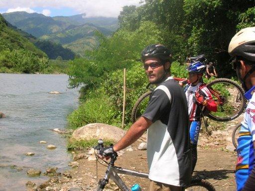 A group of mountain bikers standing by a riverbank in a lush, green landscape. One cyclist, wearing sunglasses and a black and white jersey, looks towards the camera while holding a bicycle. In the background, more cyclists are seen, with mountains and trees visible in the scenery. Cordillera Central mountain bike trail.