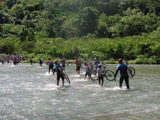 A group of cyclists wading through a river, carrying their bicycles amidst lush green vegetation and trees in the background. Bright sunlight filters through the foliage, creating a vibrant and dynamic outdoor scene. Cordillera Central mountain bike trail.