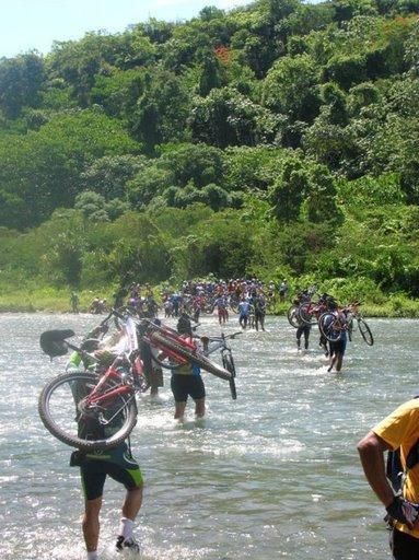 A group of people wade through a shallow river, carrying their bicycles above their heads. Lush greenery and trees line the riverbank, indicating a natural outdoor setting. The sun is shining, creating a bright and warm atmosphere. Cordillera Central mountain bike trail.