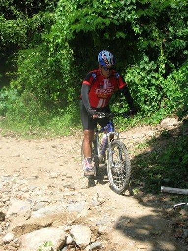 A person in a red cycling jersey and helmet is riding a mountain bike on a rocky, uneven trail surrounded by lush green vegetation. Cordillera Central mountain bike trail.