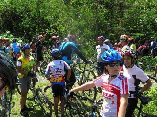 A group of mountain bikers gathered in a lush, green outdoor setting, wearing various biking gear and helmets. Some cyclists are on their bikes, while others are standing nearby, engaged in conversation or preparing for a ride. The scene captures a lively atmosphere of a biking event or gathering in nature. Cordillera Central mountain bike trail.