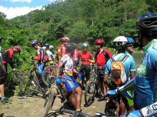 A group of cyclists in colorful jerseys and helmets gathered near a lush green area, surrounded by trees and a body of water. Some cyclists are adjusting their bikes, while others are chatting and resting. The scene is bright and sunny, showcasing a vibrant outdoor setting. Cordillera Central mountain bike trail.