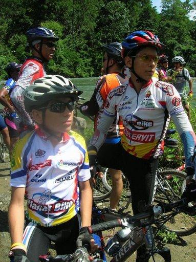 A group of cyclists wearing colorful jerseys and helmets, standing by their bikes near a river. Two cyclists in the foreground are looking off to the side, one is wearing sunglasses and a black helmet, while the other is wearing a white and red jersey. The scene is set in a lush, green outdoor environment. Cordillera Central mountain bike trail.