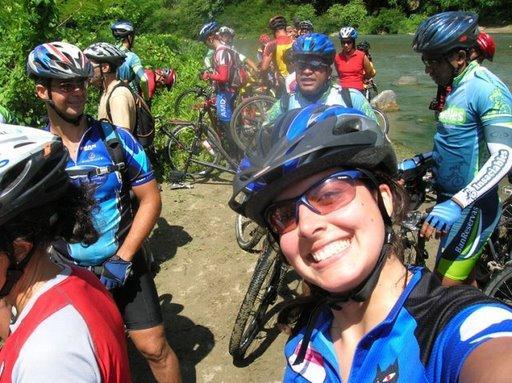 A group of cyclists in helmets and sunglasses taking a break by a river. The photo captures a smiling woman in the foreground, surrounded by other riders engaged in conversation, with lush greenery in the background. Cordillera Central mountain bike trail.