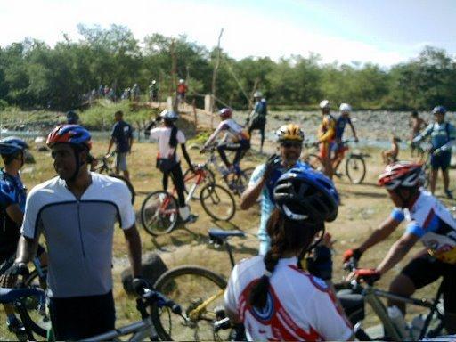 A group of cyclists dressed in colorful jerseys and helmets gathered near a river, some with their bikes. The background features trees and a bridge, indicating an outdoor cycling event or gathering. Cordillera Central mountain bike trail.