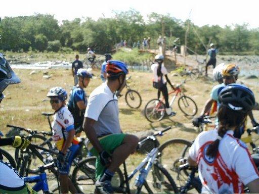 A group of cyclists in casual clothing and helmets is gathered near a body of water, with some riders on bikes and others standing beside them. In the background, a bridge and additional cyclists can be seen. The scene is sunny and features greenery in the surrounding area. Cordillera Central mountain bike trail.