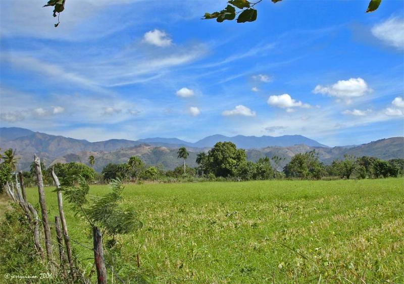 A panoramic view of a lush green field with mountains in the background under a blue sky filled with scattered clouds. The foreground features tall grasses and a few trees, while rustic wooden posts line the edge of the field, suggesting a rural landscape. Cordillera Central mountain bike trail.