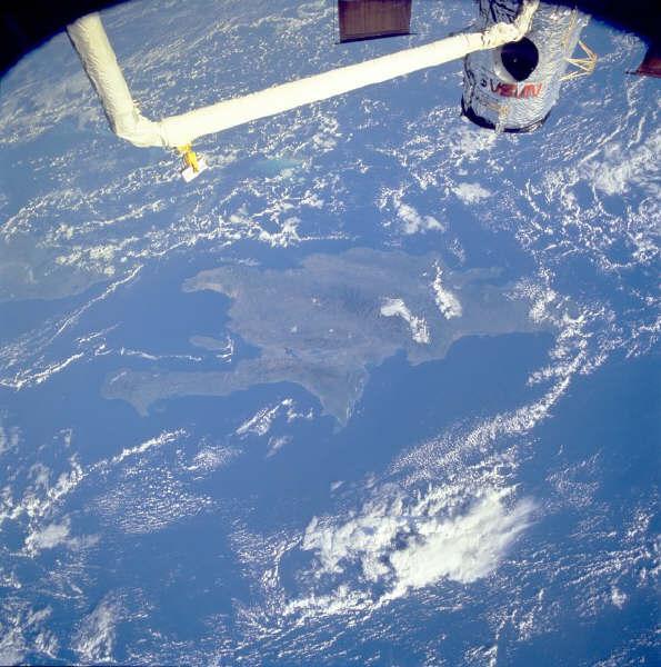 Satellite view of a landmass surrounded by ocean, with some cloud cover visible. A portion of a spacecraft's arm is seen in the foreground, extending into the image from the top left corner. Cordillera Central mountain bike trail.