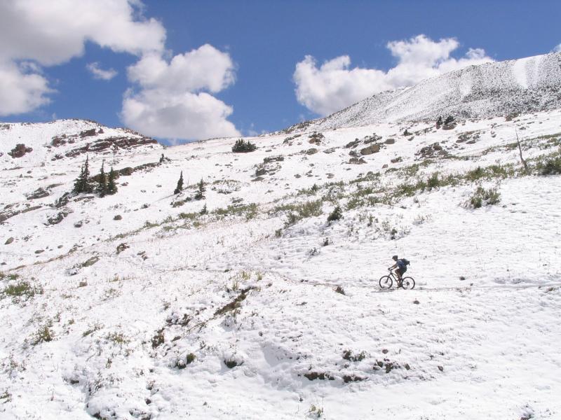 A mountain biker navigates a snow-covered landscape, surrounded by rocky hills and patches of green vegetation under a bright blue sky with fluffy white clouds. Colorado Trail: Searle Pass and Kokomo Pass (Copper Mountain to Camp Hale) mountain bike trail.