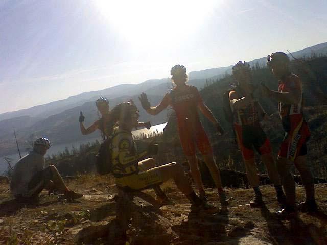 A group of six mountain bikers in cycling gear stands on a rocky outcrop, enjoying a sunny day outdoors. The background features rolling hills and a clear blue sky. The bikers are smiling and posing for the camera, with a couple of them giving thumbs up. One cyclist is seated, resting on the rocks. The scene conveys a sense of camaraderie and adventure in a natural setting. Okanagan Mountain Park mountain bike trail.