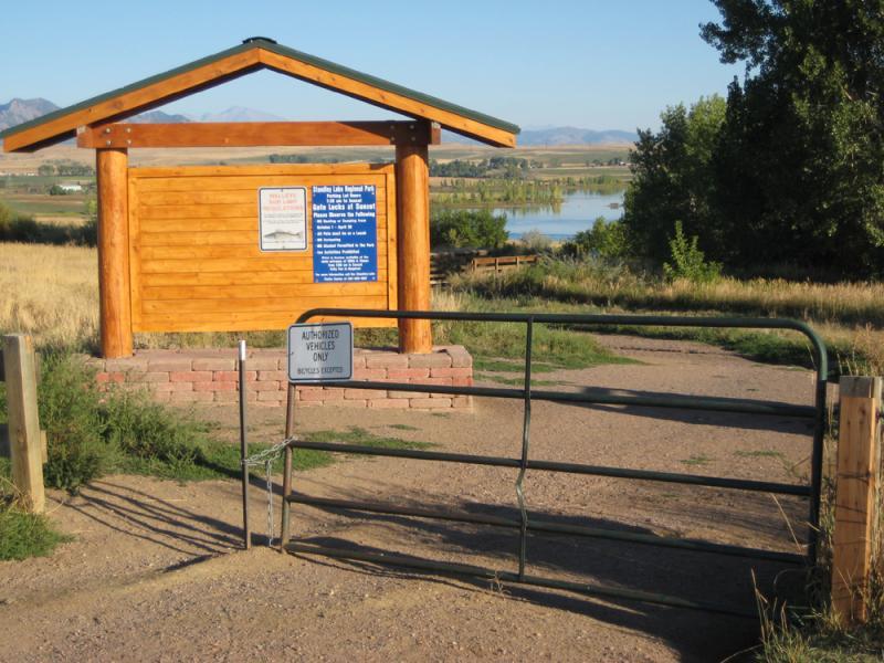 Wooden entrance sign for a recreational area near a lake, featuring a blue information board. A metal gate labeled "AUTHORIZED VEHICLES ONLY" is visible in the foreground, with grassy terrain and trees in the background. The landscape includes rolling hills and distant mountains under a clear sky. Standley Lake mountain bike trail.