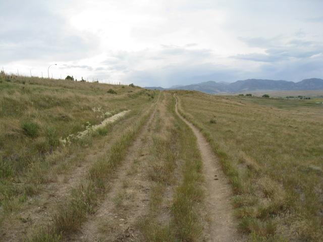 A dirt road winding through grassy terrain, flanked by gentle hills and a cloudy sky in the background. The landscape is mostly open, with sparse vegetation. Standley Lake mountain bike trail.