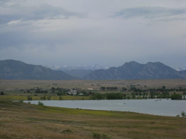 A scenic view of a landscape featuring rolling hills and mountains in the background under a cloudy sky. A calm body of water is in the foreground, surrounded by grassy fields and sparse trees. The distant mountains are partially obscured by clouds, hinting at snow-capped peaks. Standley Lake mountain bike trail.