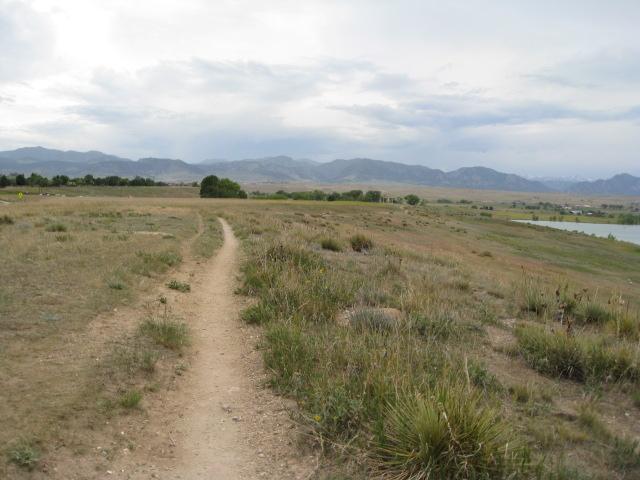 A winding dirt path leads through a grassy landscape toward a distant lake, with mountains visible in the background under a cloudy sky. Standley Lake mountain bike trail.