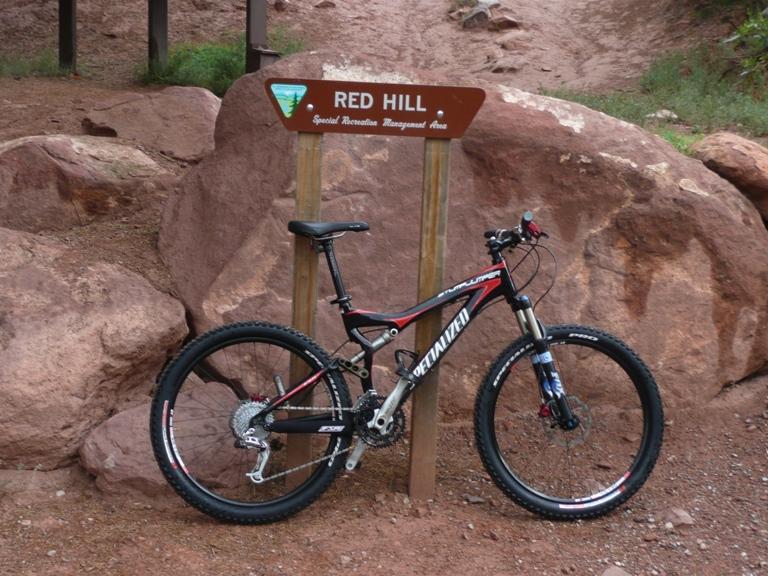A mountain bike leaning against a sign that reads "RED HILL" in a Special Recreation Management Area, surrounded by rocky terrain and greenery. Red Hill mountain bike trail.