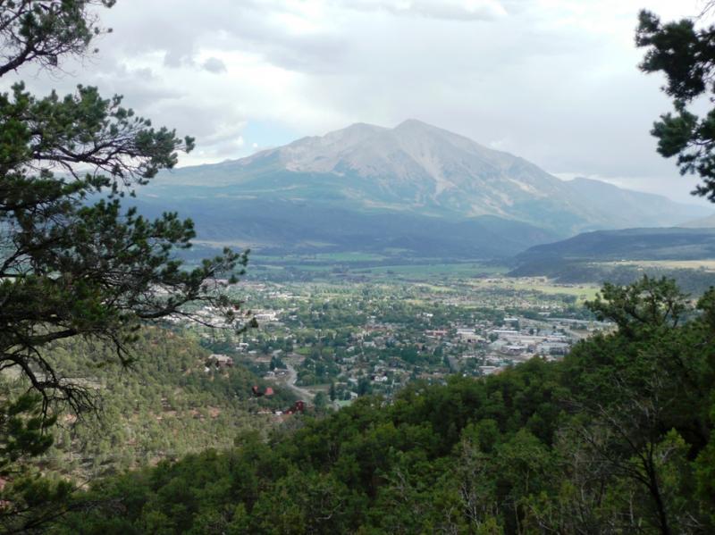A panoramic view of a mountainous landscape with lush green forests in the foreground, leading to a valley and a small town below. A prominent mountain peak rises in the background under a partly cloudy sky, creating a serene and picturesque natural setting. Red Hill mountain bike trail.