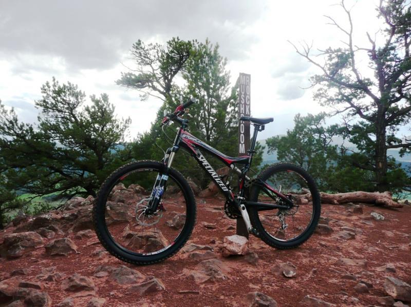 A mountain bike parked on rocky terrain with a trail sign in the background, surrounded by trees and cloudy skies. Red Hill mountain bike trail.