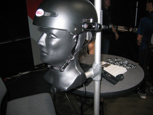 A black Bell helmet displayed on a mannequin head, attached to a metal stand beside a table with promotional materials. The background is dark, and people can be seen in the distance.