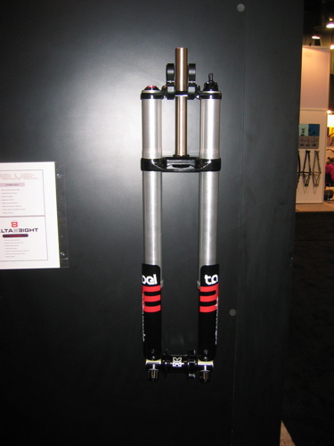 Mountain bike suspension fork displayed on a black wall. The fork features two silver aluminum stanchions with black and red branded lower legs. It includes adjustment knobs at the top and a sleek design suitable for off-road biking. A nearby sign provides additional information about the product.