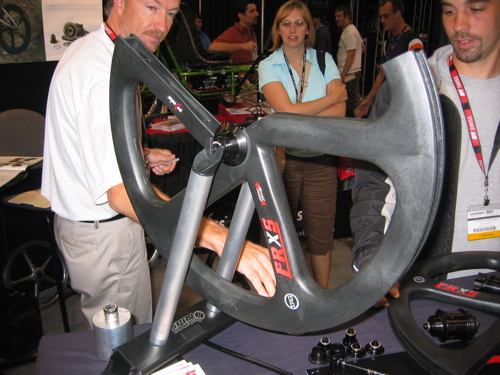 A group of people at a trade show examining a large, modern bicycle wheel displayed on a stand. The wheel features a sleek design with a carbon fiber look, and one person is interacting with it while others watch. The background includes additional cycling equipment and attendees engaged in conversation.