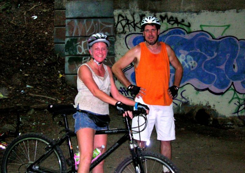 Two individuals standing next to a black bicycle in a graffiti-covered outdoor area. One person, wearing a white tank top and denim shorts, is smiling and wearing a helmet. The other person, dressed in an orange tank top and white shorts, has a relaxed pose with hands on hips. The setting appears to be a shaded spot, surrounded by greenery and urban art on the wall behind them. Wissahickon Valley Park mountain bike trail.