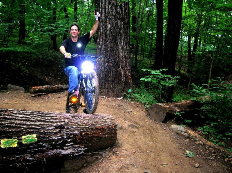 A mountain biker riding over a fallen log in a wooded area, raising one hand in celebration. The bike is equipped with a front light, illuminating the path ahead. Trees and lush greenery surround the scene, suggesting an adventurous outdoor environment. Wissahickon Valley Park mountain bike trail.