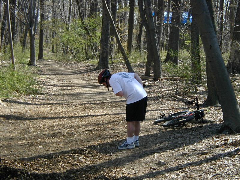 A person wearing a helmet and a white t-shirt leans over to examine the ground on a dirt trail in a wooded area. A bicycle lies on its side nearby, surrounded by trees and scattered leaves, indicating a recreational outdoor activity. Wissahickon Valley Park mountain bike trail.