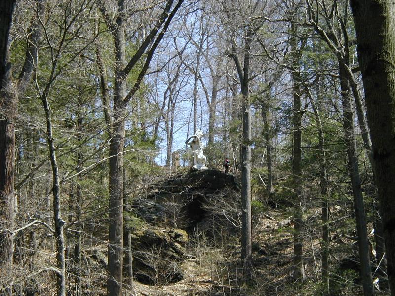 A white statue perched on a rocky outcrop surrounded by bare trees and greenery in a forest setting, with a person visible in the background. The sky is clear and blue, indicating a sunny day. Wissahickon Valley Park mountain bike trail.