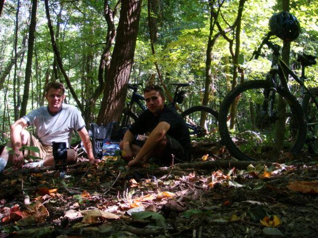 Two young men sitting on the forest floor surrounded by trees, with their mountain bikes propped up beside them. The ground is covered in leaves, and sunlight filters through the trees, casting warm tones on the scene. Great Seal State Park mountain bike trail.