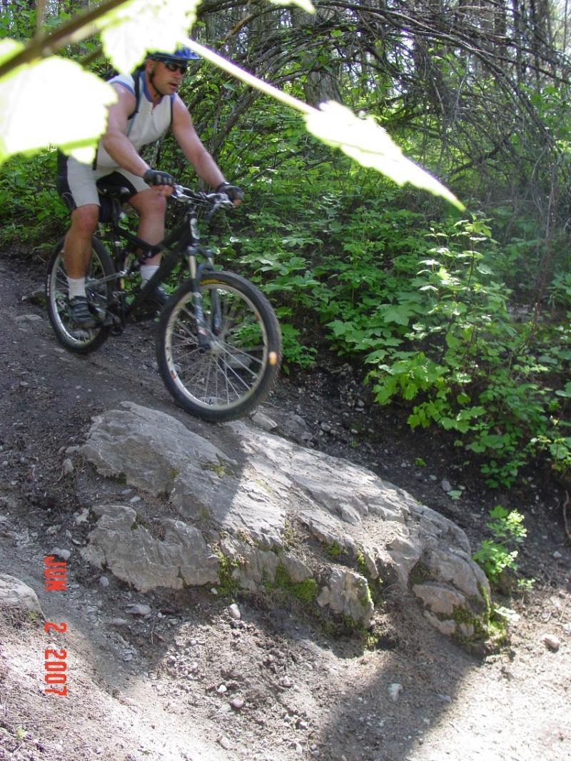 A mountain biker navigating a rocky trail surrounded by lush greenery. The cyclist is wearing a helmet and riding gear, focused on maintaining balance as they approach a large rock on the path. Sunlight filters through the leaves, creating a vibrant outdoor scene. Spencer Mountain mountain bike trail.