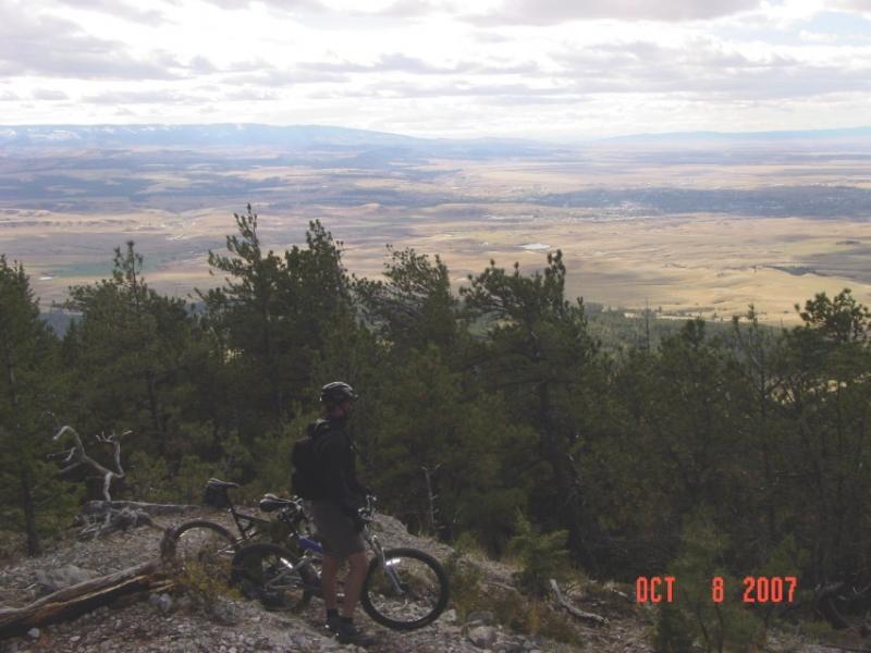 A person stands with a mountain bike on a rocky outcrop, overlooking a vast valley and distant mountains. The scene is framed by pine trees and features a cloudy sky. The date "OCT 8 2007" is visible in the lower right corner. Limekiln mountain bike trail.