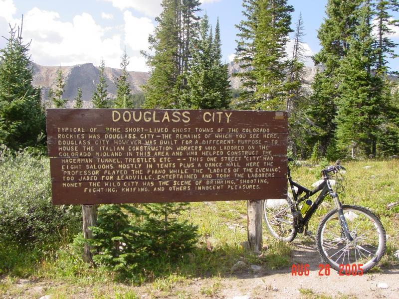 A weathered wooden sign labeled "Douglass City" stands surrounded by tall trees in a mountainous area. The sign describes Douglass City as a short-lived ghost town in the Colorado Rockies, highlighting its history as a site for Italian construction workers involved in building the Colorado Midland. A bicycle is leaning against the sign, and the date "AUG 20 2005" is visible at the bottom. The background features green foliage and distant mountains under a clear sky. Hagerman Tunnel mountain bike trail.