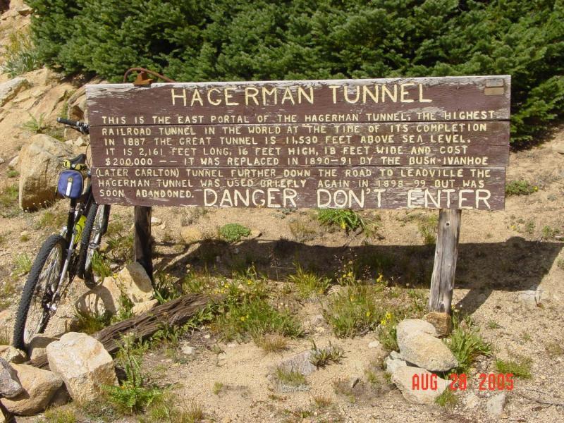 A weathered wooden sign post detailing information about the Hagerman Tunnel, located at its east portal. The sign includes historical facts about the tunnel's height, length, and construction cost, along with a cautionary note stating "DANGER DON'T ENTER." A parked bicycle stands nearby, with a backdrop of greenery and rocky terrain. The date "AUG 20, 2005" is visible in the lower right corner of the image. Hagerman Tunnel mountain bike trail.