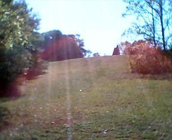 A grassy hillside under a clear blue sky, with trees on both sides and a few colorful autumn leaves visible. The sunlight creates a lens flare effect on the image. Athens Regional Park Trail mountain bike trail.