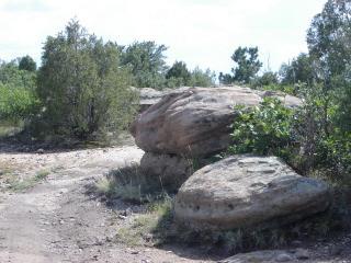 A rocky landscape featuring large boulders alongside a dirt path, surrounded by low shrubs and trees under a partly cloudy sky. Hidden Mesa mountain bike trail.