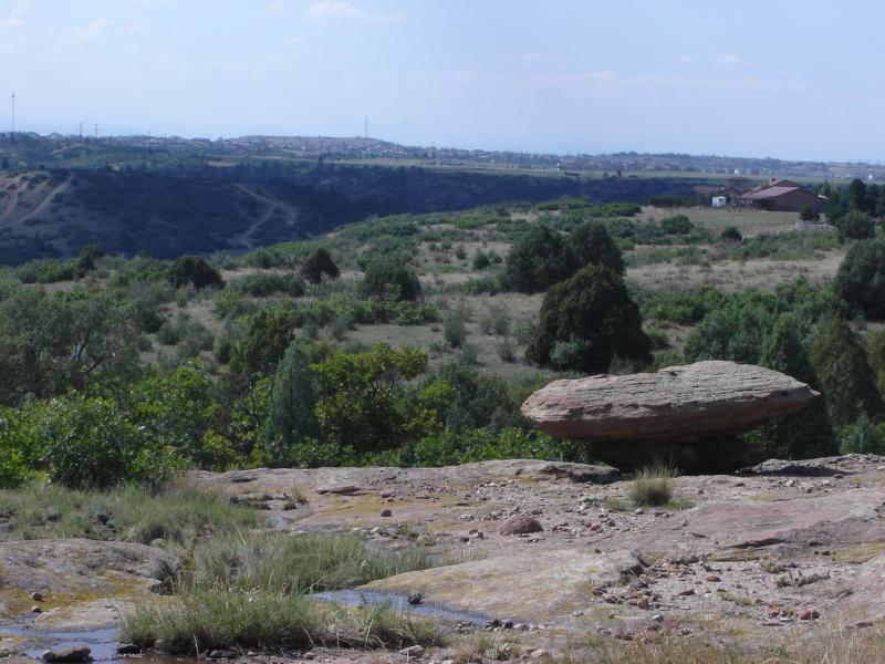 A scenic view of rolling hills and a plateau featuring a large, flat rock formation in the foreground. The landscape is lush with greenery, including various shrubs and trees, while distant hills and a farmstead can be seen in the background under a partially cloudy sky. Hidden Mesa mountain bike trail.