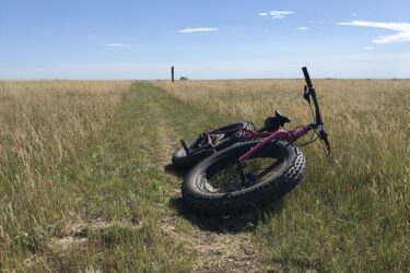 A purple fat tire bike resting on a grassy pathway that stretches through an open field, with a clear blue sky overhead and a distant vertical post visible along the trail. Long X mountain bike trail.