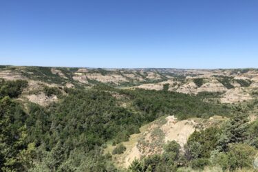 A panoramic view of rolling hills and valleys, featuring a mix of lush green vegetation and exposed rock formations under a clear blue sky. The landscape showcases natural geological features and is filled with sparse trees and shrubs. Long X mountain bike trail.
