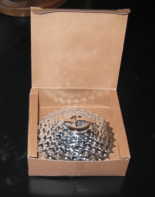 A shiny bicycle cassette resting inside an open cardboard box, showcasing its intricate gear teeth and central sprocket. The box has a light brown color and is partially closed, with the cassette visible from above. The background is a dark surface, emphasizing the metallic shine of the cassette.