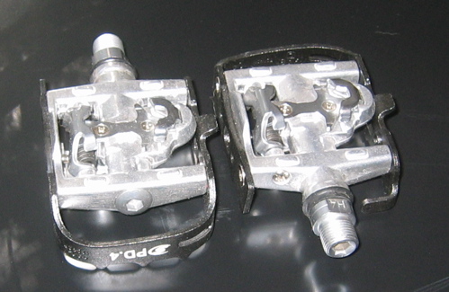 Close-up view of two bicycle pedals, showcasing their metallic design and adjustable cleat system. The pedals are silver with black accents, featuring the brand logo on one side and a threaded spindle for attachment to a bike.