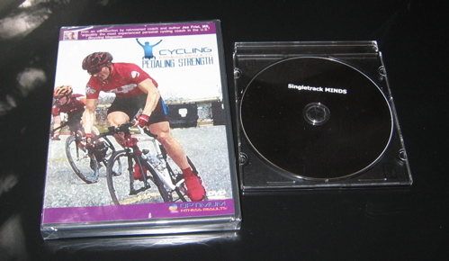 A DVD case for a cycling workout titled "Cycling Pedaling Strength," featuring an image of a cyclist in action on the cover. Next to it, a black DVD disc is placed in a clear case. The disc is labeled "Singletrack MENDS."