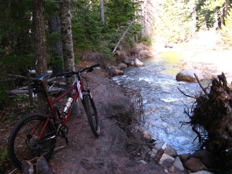 A mountain bike rests on a dirt path beside a flowing stream, surrounded by lush green trees and rocky terrain. Sunlight filters through the foliage, illuminating the scene. E Fork Hood River mountain bike trail.