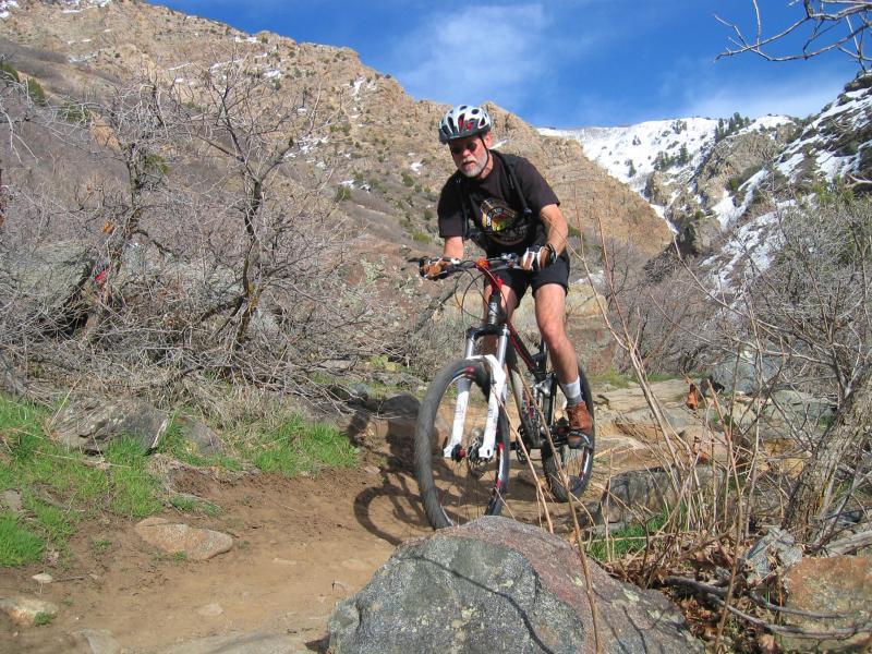 A mountain biker navigating a rocky trail in a mountainous landscape. The rider, wearing a helmet and casual cycling attire, is focused on maneuvering his bike around large boulders and through sparse vegetation. Snow-capped peaks are visible in the background under a partly cloudy sky. Bonneville Shoreline Trail - Ogden Section mountain bike trail.