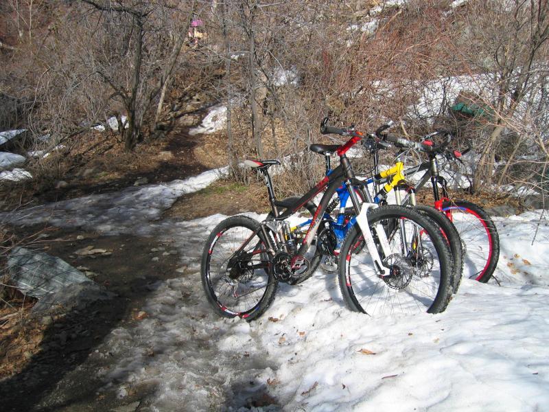 Three mountain bikes parked near a snowy trail surrounded by bare trees and underbrush on a winter day. Bonneville Shoreline Trail - Ogden Section mountain bike trail.