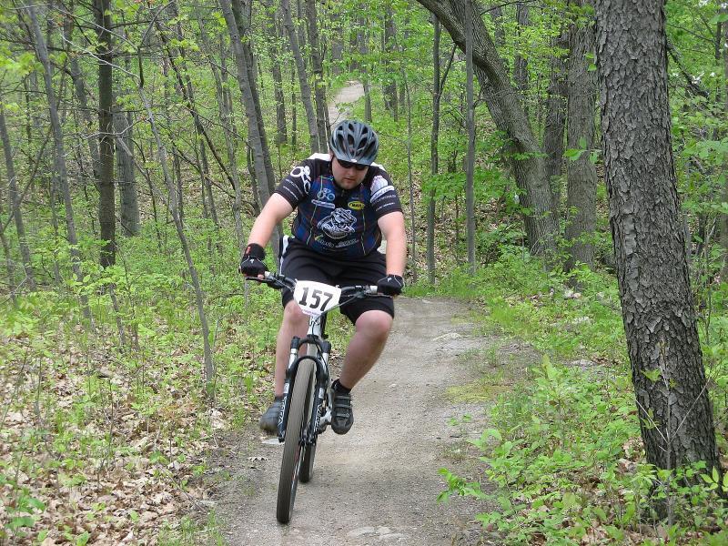 A cyclist in a black helmet and a blue and black jersey rides a mountain bike along a dirt path in a lush, green forest. The rider is focused and balanced, with trees and foliage visible on either side. The image captures the essence of outdoor adventure and mountain biking. Addison Oaks mountain bike trail.