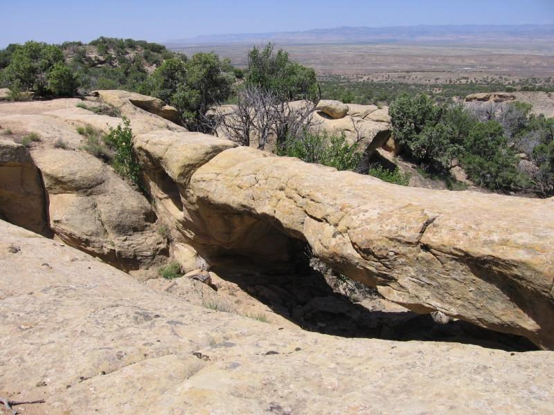 A rocky landscape featuring a natural stone arch, surrounded by sparse vegetation and distant hills under a clear blue sky. The sun highlights the textures of the sandstone and the greenery along the edges. Westwater Mesa / Overlook mountain bike trail.